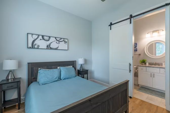 Bright modern bedroom with light-blue bedding on a dark wood bed, matching nightstands and lamps, abstract wall art, and a sliding barn door open to a white en-suite bathroom with round mirror and vanity.