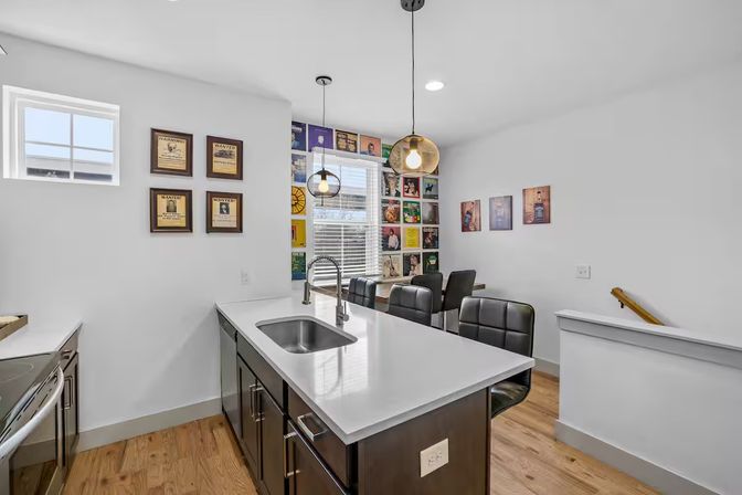 Bright modern open-concept kitchen with white quartz island and undermount sink, dark wood cabinets, black leather bar stools at a breakfast bar, globe pendant lights, hardwood floors, large window with blinds, and a colorful vinyl record gallery wall in the dining nook.