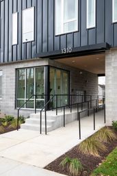 Modern urban apartment entrance numbered 1310: concrete steps and accessible ramp leading to glass entry doors beneath black metal siding with simple sidewalk landscaping.