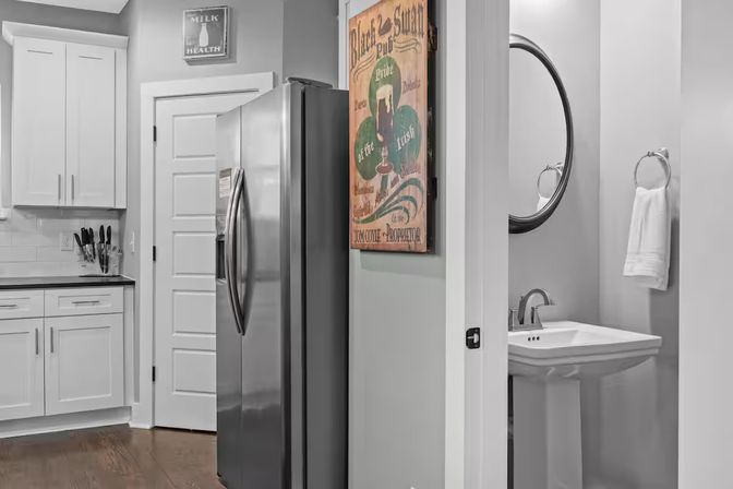 Modern home kitchen with stainless steel refrigerator beside white shaker cabinets and knife block, hardwood floors, decorative wall sign, and an open doorway to a powder room with pedestal sink and round mirror.
