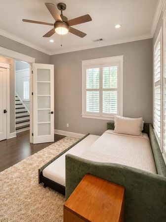 Cozy, bright guest bedroom with green upholstered daybed and pull-out trundle, white plantation shutters, ceiling fan, plush rug, and hardwood floors.