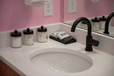 Cheerful pink bathroom vanity with white countertop and oval undermount sink, matte black faucet, glass jars holding cotton pads and swabs, and a wall mirror.