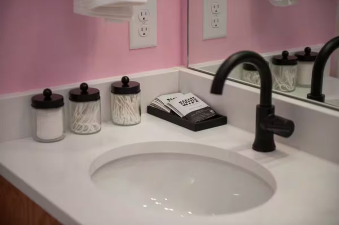 Cheerful pink bathroom vanity with white countertop and oval undermount sink, matte black faucet, glass jars holding cotton pads and swabs, and a wall mirror.