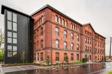 Large historic red brick industrial building with rows of arched windows, a modern black metal glass stair tower at one end, landscaped curb, and wet parking lot reflecting the facade on an overcast day.