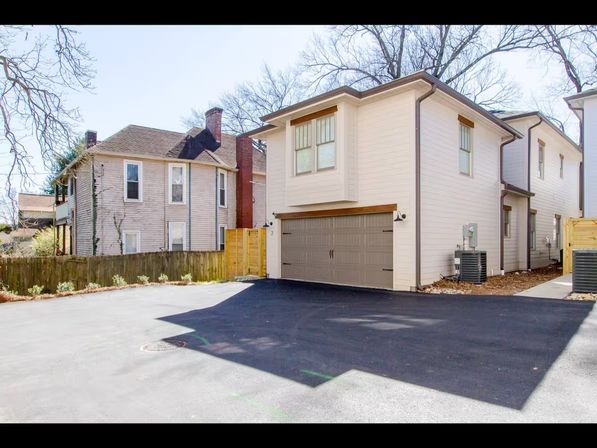 Sunlit beige two-story house with two-car garage, smooth paved driveway, wooden privacy fence and older neighboring home framed by bare trees