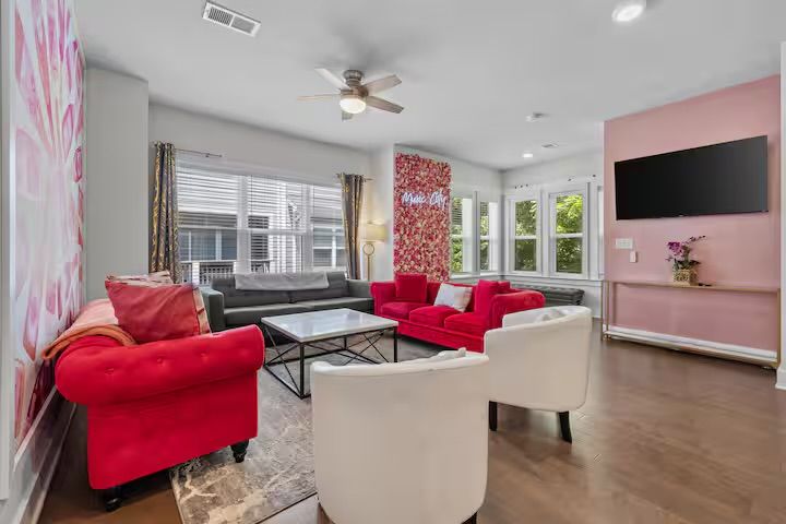 Bright modern living room interior with bold red velvet sofas, white accent chairs, gray sectional, glass coffee table, floral accent wall, bay windows and hardwood floors — playful, light-filled design.