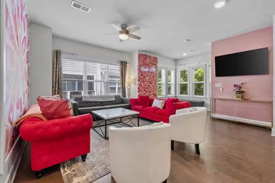 Bright modern living room interior with bold red velvet sofas, white accent chairs, gray sectional, glass coffee table, floral accent wall, bay windows and hardwood floors — playful, light-filled design.