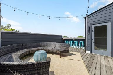 Sunny rooftop deck with a curved dark wicker sectional circling a round blue ottoman, string lights overhead, turquoise bar stools along the railing, and weathered wooden decking under a clear blue sky.