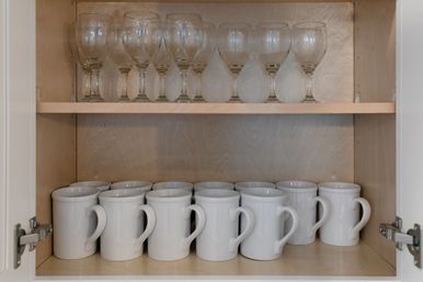 Organized kitchen cabinet with clear wine glasses on the top shelf and neatly lined white ceramic mugs on the bottom shelf against a light wood interior.