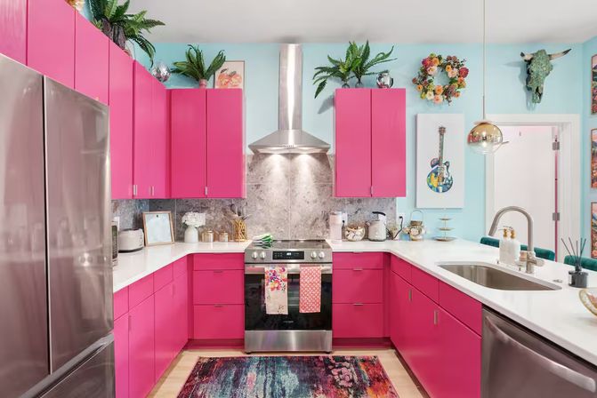 Cheerful modern kitchen with hot-pink cabinets, stainless steel appliances, marble backsplash and white countertops, turquoise walls, potted plants and a colorful area rug.