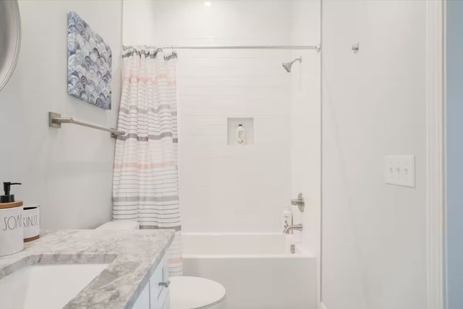 Bright modern residential bathroom interior with white bathtub-shower, pastel striped shower curtain, marble vanity countertop and brushed nickel fixtures