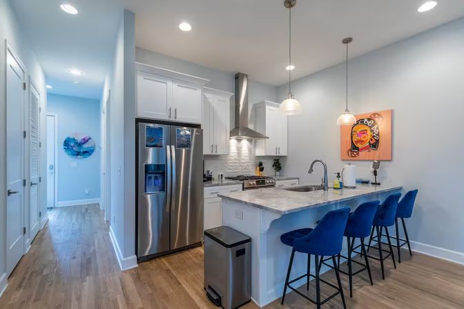 Modern open-concept kitchen with white cabinetry, marble island, stainless steel fridge and range, four blue velvet bar stools, pendant lights and hardwood floors
