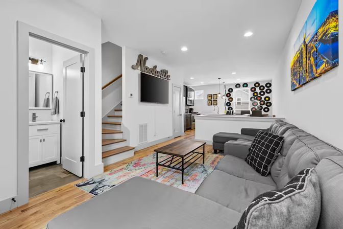 Bright modern Nashville rental living room — cozy gray sectional with patterned pillows, wooden coffee table on a colorful rug, wall-mounted TV under a "Nashville" sign, staircase, and open kitchen with bar and vinyl record wall art.
