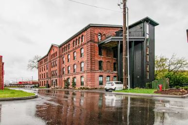 Historic red brick industrial building with a modern black metal stair/elevator tower, parked car and rail cars in the background on a rainy day with wet pavement reflections.