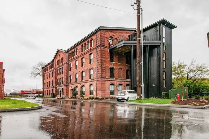 Historic red brick industrial building with a modern black metal stair/elevator tower, parked car and rail cars in the background on a rainy day with wet pavement reflections.