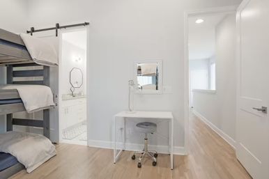 Bright modern bedroom in a home interior with stacked wooden bunk beds, compact white desk and rolling stool, hardwood floors, and a sliding barn-door to an en-suite bathroom.