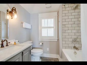 Bright modern residential bathroom — marble vanity with bronze faucet, wall sconce and towel, toilet beneath shuttered window, and bathtub with gray-grout subway tile shower.