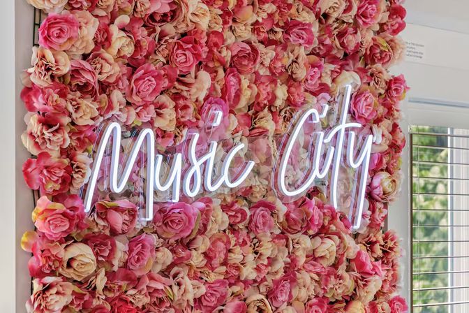 White neon script sign mounted on a wall covered in dense pink and cream roses, creating a bright floral photo backdrop