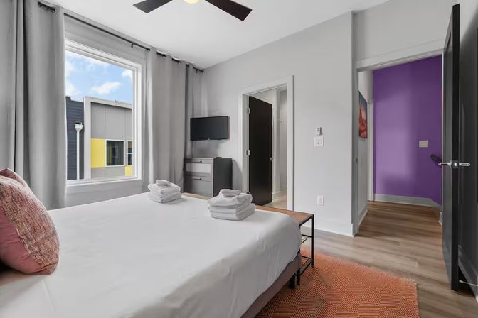 Bright, modern urban apartment bedroom with king bed, folded towels, wall-mounted TV, large window showing a yellow-accent building, gray curtains and hardwood floors