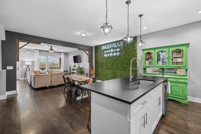 Nashville open-concept kitchen and living room with large island and black countertop, pendant lights, green moss accent wall with neon “Nashville” sign, vintage green hutch, dining table and hardwood floors.