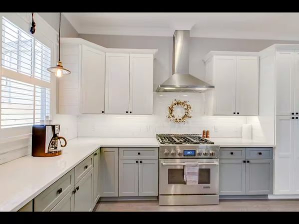 Bright modern kitchen with white upper cabinets and gray lower cabinets, stainless steel gas range and hood, white quartz countertops, pendant light, coffee maker and wreath above stove