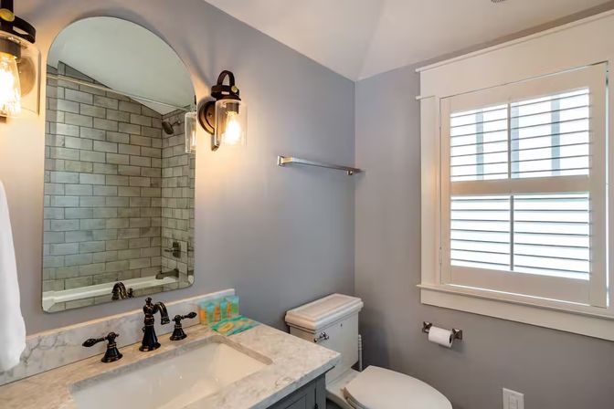 Bright modern bathroom with marble countertop and undermount sink, arched mirror, oil-rubbed bronze faucets, glass wall sconces, subway-tile shower and white plantation shutters.