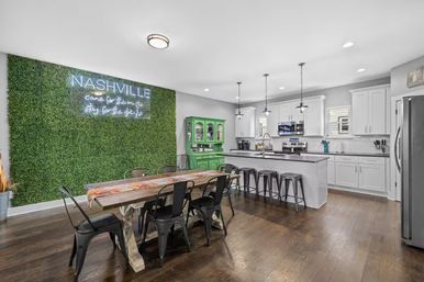 Open-concept Nashville kitchen and dining area with a green hedge wall and neon sign, farmhouse table with black metal chairs, white cabinets, island with bar stools and stainless appliances on dark hardwood floors.