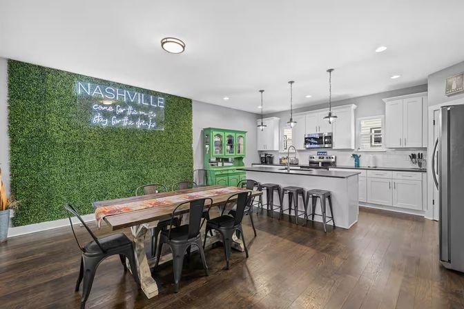Open-concept Nashville kitchen and dining area with a green hedge wall and neon sign, farmhouse table with black metal chairs, white cabinets, island with bar stools and stainless appliances on dark hardwood floors.