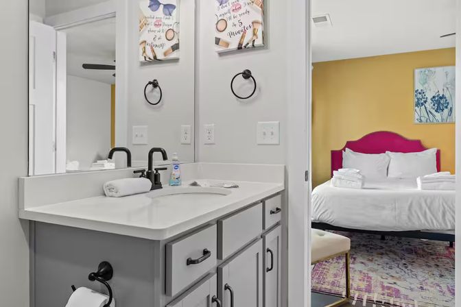 Modern bathroom vanity with white countertop, matte black faucet and rolled towel beside a mirror, view into a bedroom with a fuchsia headboard, yellow accent wall and neatly made white bed with folded towels.
