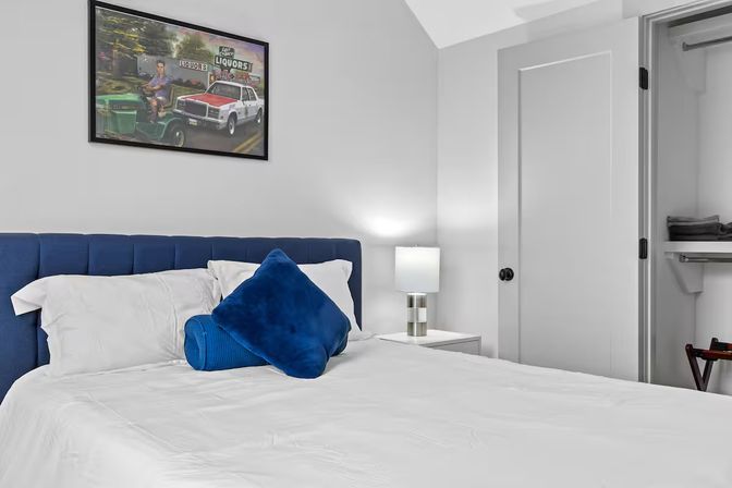 Crisp modern bedroom with cobalt blue tufted headboard and pillows, white linens, bedside lamp, framed car artwork on wall and open closet