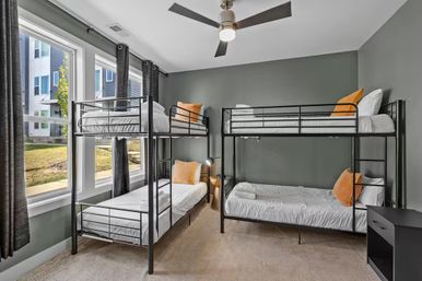 Bright modern shared bedroom in an urban apartment with two black metal bunk beds (four beds), gray walls, large windows with natural light, white bedding, orange accent pillows and a ceiling fan.