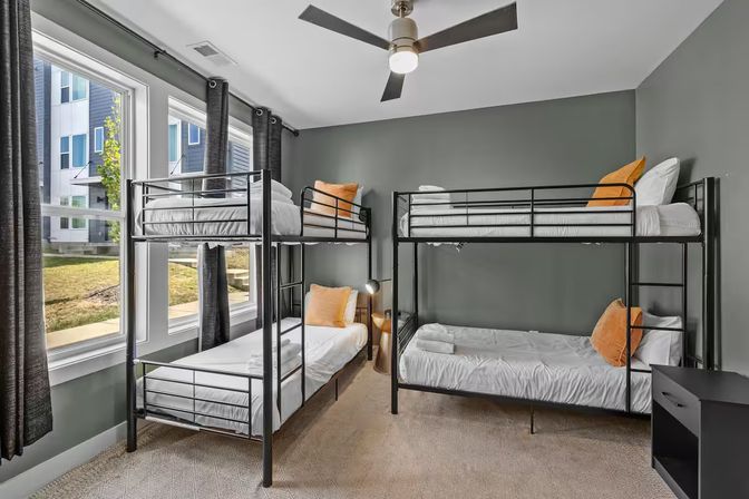 Bright modern shared bedroom in an urban apartment with two black metal bunk beds (four beds), gray walls, large windows with natural light, white bedding, orange accent pillows and a ceiling fan.