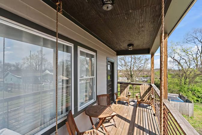 Sunlit covered wooden porch with bistro table, Adirondack chairs and hanging rope swing, overlooking a suburban backyard and tree-lined view.