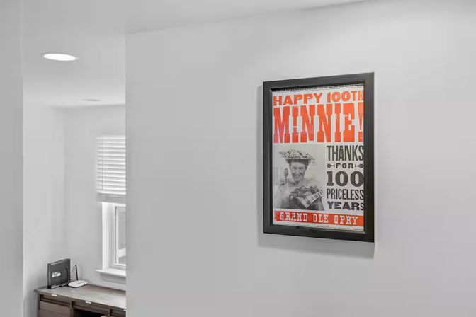 Framed vintage-style 'Happy 100th' poster with bold orange lettering on a light-gray hallway wall above a small home office nook with desk, router and window blinds