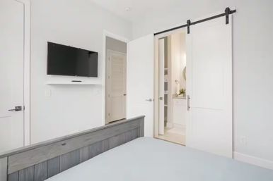 Bright modern bedroom with gray wood bedframe, wall-mounted flat-screen TV, and white sliding barn door revealing an en-suite bathroom in a neutral, minimalist decor.