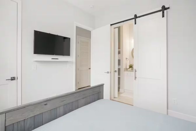Bright modern bedroom with gray wood bedframe, wall-mounted flat-screen TV, and white sliding barn door revealing an en-suite bathroom in a neutral, minimalist decor.