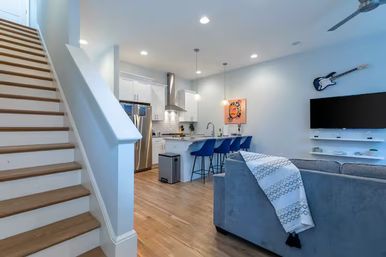 Bright modern open-concept living room and kitchen with hardwood floors, white island and three blue bar stools, stainless appliances, gray sofa with patterned throw, wall-mounted TV and decorative electric guitar.