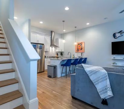 Bright modern open-concept living room and kitchen with hardwood floors, white island and three blue bar stools, stainless appliances, gray sofa with patterned throw, wall-mounted TV and decorative electric guitar.