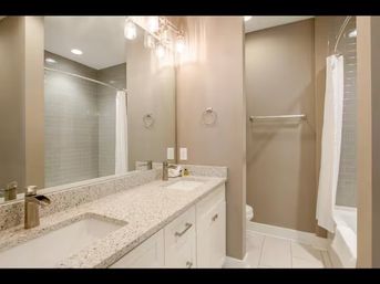 Modern neutral bathroom with double vanity and undermount sinks in a speckled granite countertop, white cabinets, large mirror, pendant lighting, towel rack, and a tiled shower with white curtain.