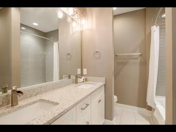 Modern neutral bathroom with double vanity and undermount sinks in a speckled granite countertop, white cabinets, large mirror, pendant lighting, towel rack, and a tiled shower with white curtain.