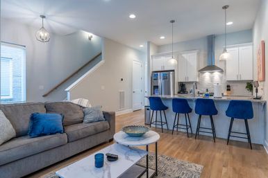 Bright modern open-concept living room and kitchen with a gray sofa and blue throw pillows, marble coffee table, hardwood floors, a breakfast island with four blue bar stools, stainless steel appliances, white cabinets and pendant lights.