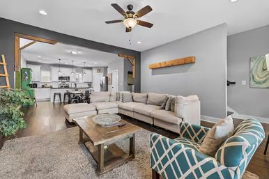 Open-concept modern farmhouse living room with beige L-shaped sectional, wooden coffee table, teal patterned armchair, ceiling fan, exposed wood beams and view into a bright white kitchen on hardwood floors.