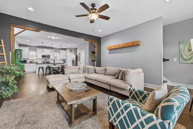 Open-concept modern farmhouse living room with beige L-shaped sectional, wooden coffee table, teal patterned armchair, ceiling fan, exposed wood beams and view into a bright white kitchen on hardwood floors.