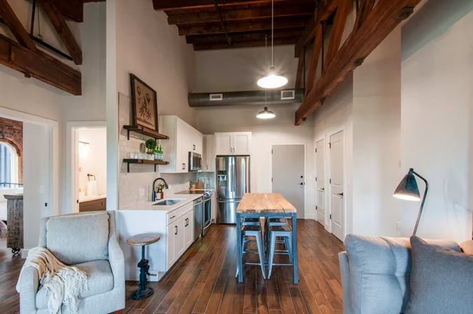 Open-concept urban loft kitchen with exposed wooden beams and high ceilings, stainless steel appliances, white cabinets, wooden island with metal stools, hardwood floors, pendant lights and cozy seating.