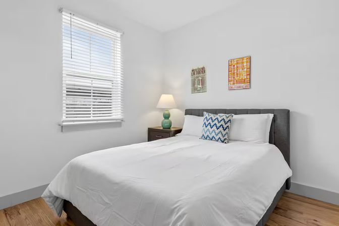 Bright minimalist bedroom with white bedding, gray upholstered headboard, blue chevron accent pillow, green bedside lamp on a dark nightstand, hardwood floor and window blinds letting in natural light.