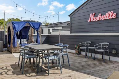 Rooftop deck with wooden dining table and gray chairs, wicker canopy daybed with blue curtains, string lights overhead, and pink "Nashville" neon sign on gray siding.