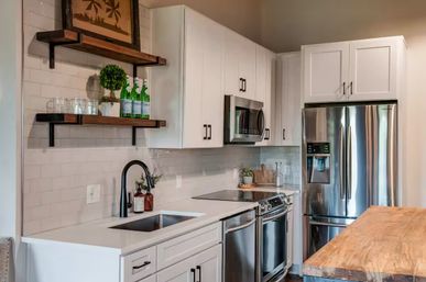 Bright modern farmhouse kitchen with white shaker cabinets, stainless-steel refrigerator and range, subway tile backsplash, black gooseneck faucet, open wood shelves and a butcher-block island.