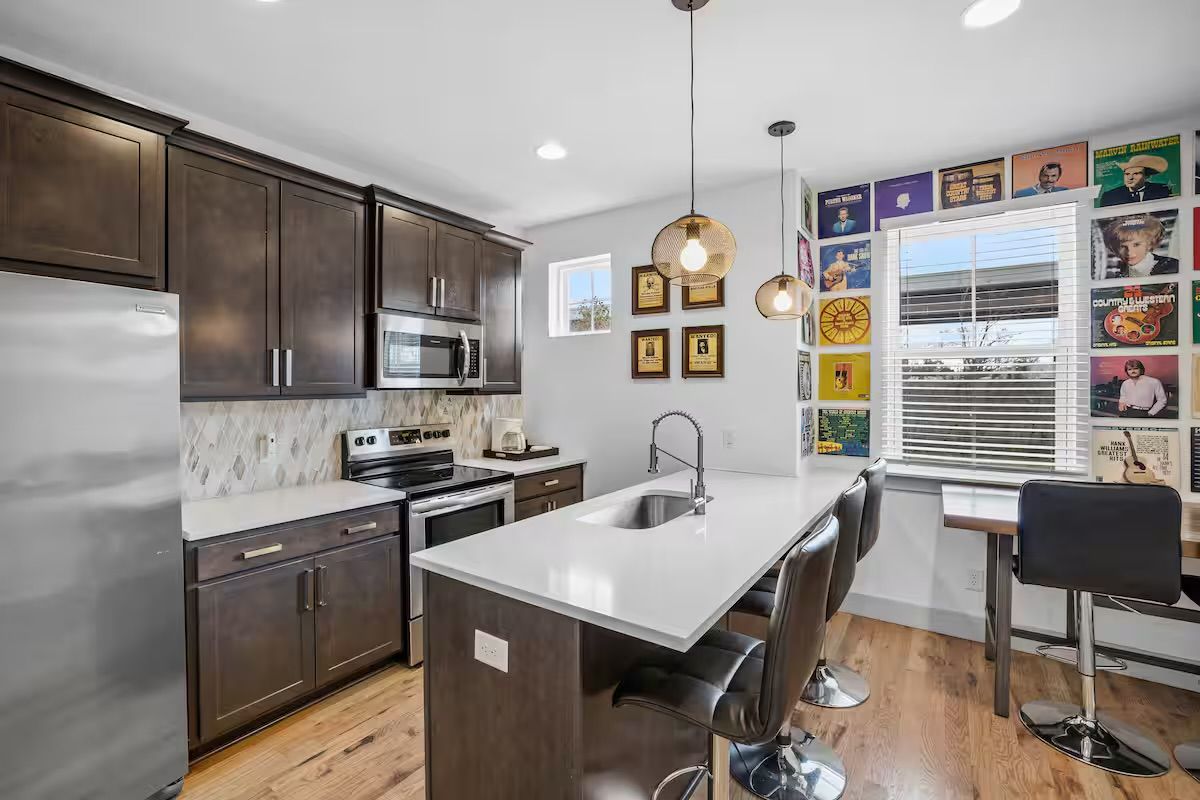 Bright modern kitchen with white quartz island and undermount sink, dark wood cabinets, stainless steel appliances, three black bar stools under woven pendant lights, and a colorful vinyl record cover gallery wall by a window.