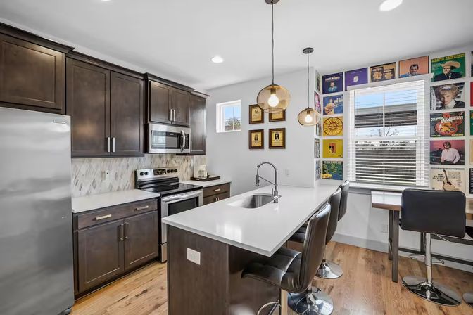 Bright modern kitchen with white quartz island and undermount sink, dark wood cabinets, stainless steel appliances, three black bar stools under woven pendant lights, and a colorful vinyl record cover gallery wall by a window.