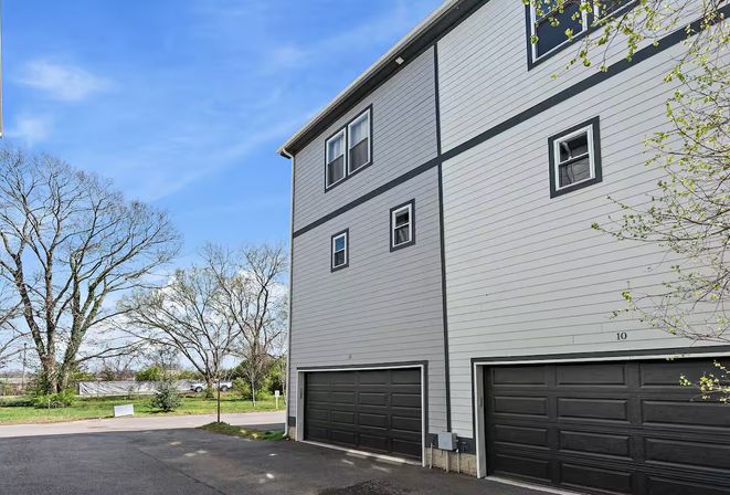 Gray three-story townhouse exterior with black garage doors, small windows, budding trees and an open suburban street under a bright blue sky